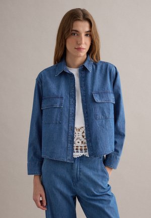 Jeune femme debout portant une veste et un pantalon en denim bleu avec un haut en dentelle blanche partiellement visible en dessous, fond neutre.