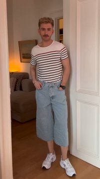 Moškp z brado, oblečen v prugasto majico, široke jeans kratke hlače, bele športne copate in uro, stoji v obliki rahlo osvetljene odprtine v prostoru.
