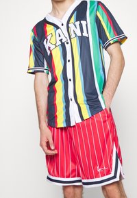 Veelgekleurd gestreept shirt met "KANI"-logo in marineblauw, geel, groen en rood; gecombineerd met rood met fijne strepen korte broek en witte randaccenten.