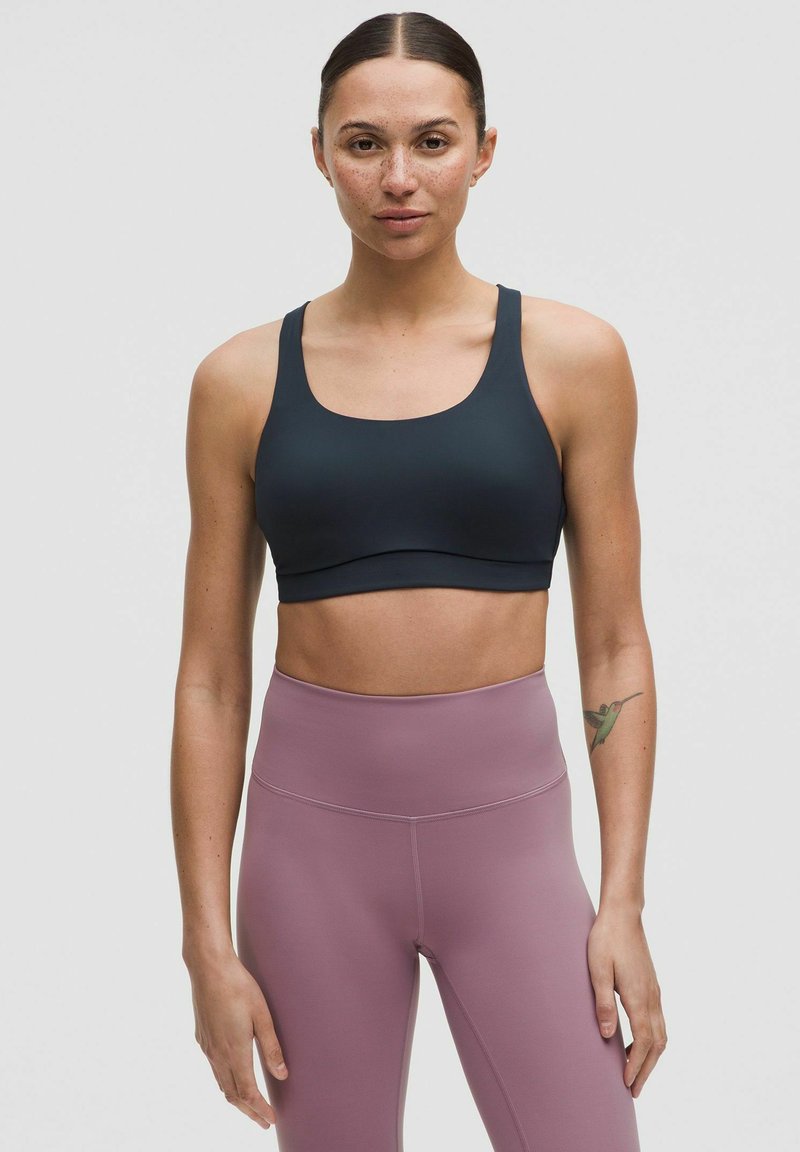 Sujetador deportivo azul marino con un escote redondeado y tirantes anchos, combinado con leggings de cintura alta en un suave tono rosa claro con una textura lisa.