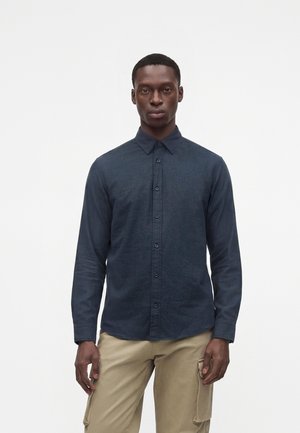 Homme portant une chemise bleue foncée boutonnée et un pantalon cargo beige, debout devant un fond blanc uni.