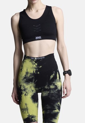 Atletisch persoon draagt een zwarte sportbeha en hoogtailleshoge zwarte en gele tie-dye legging met een fitnesshorloge om de linkerpols.