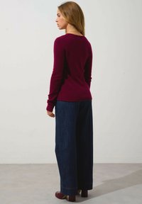 Burgunderfarbener gestrickter Pullover mit langen Ärmeln, kombiniert mit weit geschnittenen Jeanshosen und roten Plateauschuhen, von hinten betrachtet.