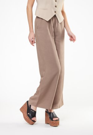 Donna che indossa pantaloni beige a gamba larga, un gilet beige e sandali con zeppa piattaforma neri con suola in sughero.
