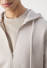 Sudadera con cremallera en beige claro, con capucha, hecha de un tejido suave. Presenta una cremallera lisa y un cuello acanalado, superpuesta sobre un cuello alto.
