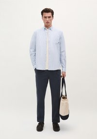 Camicia a righe blu chiaro e bianca con bottoni sopra una maglietta color crema, abbinata a pantaloni blu scuro. Il modello tiene una borsa marrone e blu.
