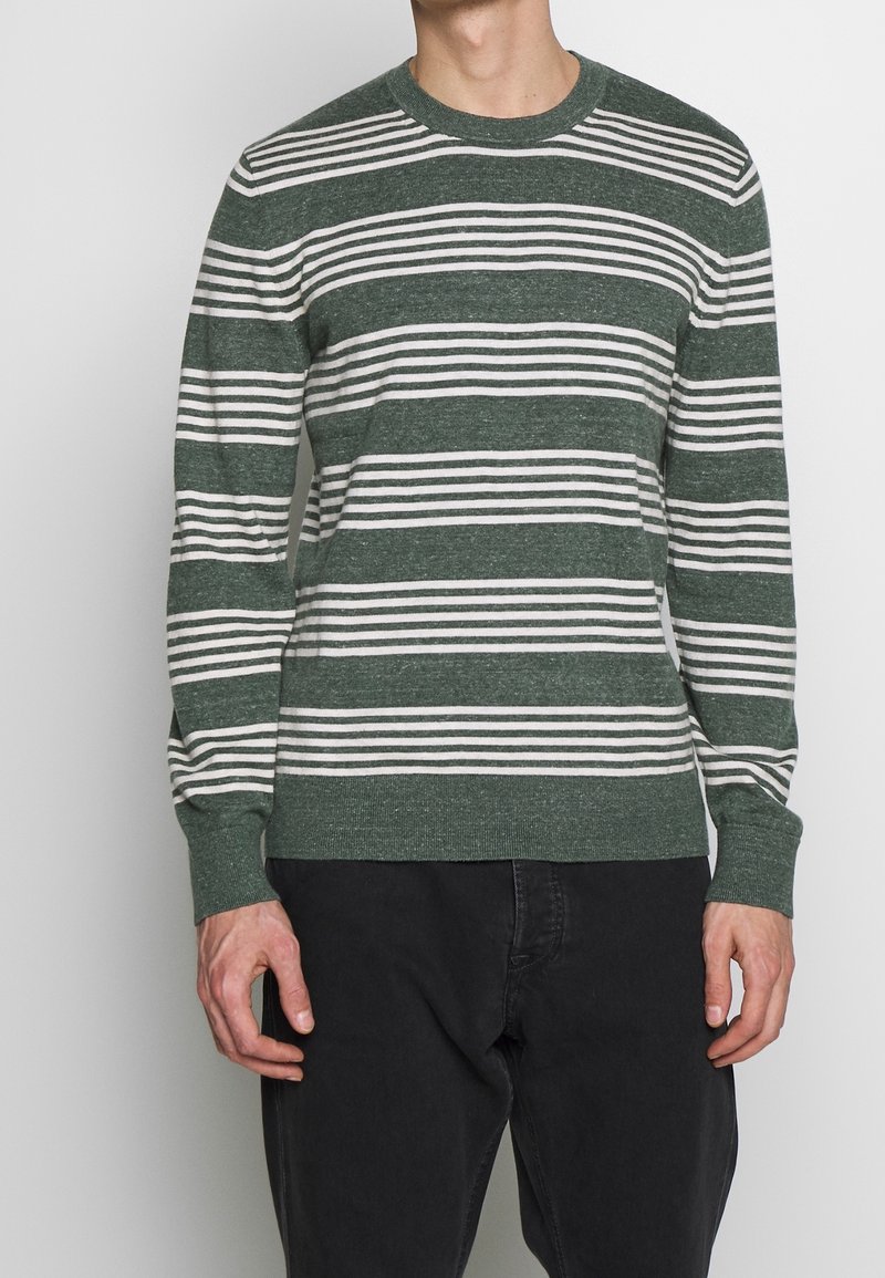 Homme portant un pull à manches longues rayé horizontalement vert et blanc et un pantalon noir, debout devant un fond clair uni.