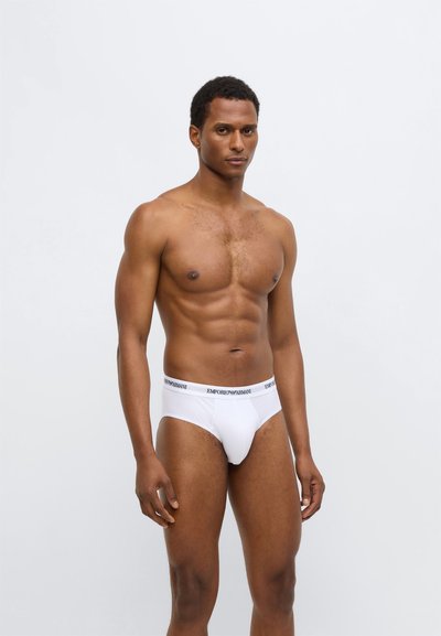 Culottes en coton blanc avec une ceinture noire portant le logo "Emporio Armani". Texture lisse avec une coupe classique, design ajusté et couverture minimale.