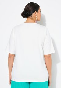 Camiseta blanca de manga corta con cuello redondo y textura suave, llevada con pantalones turquesa, vista desde la parte de atrás.
