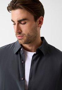 Donkergrijs shirt met een gladde textuur, klassieke kraag en paarse knopen, gelaagd over een wit shirt dat gedeeltelijk zichtbaar is.