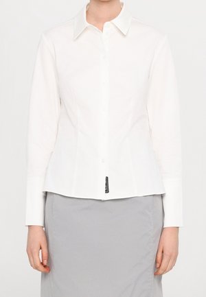 Chemise blanche à manches longues avec boutons et col, portée avec une jupe gris clair, montrée du cou aux hanches.