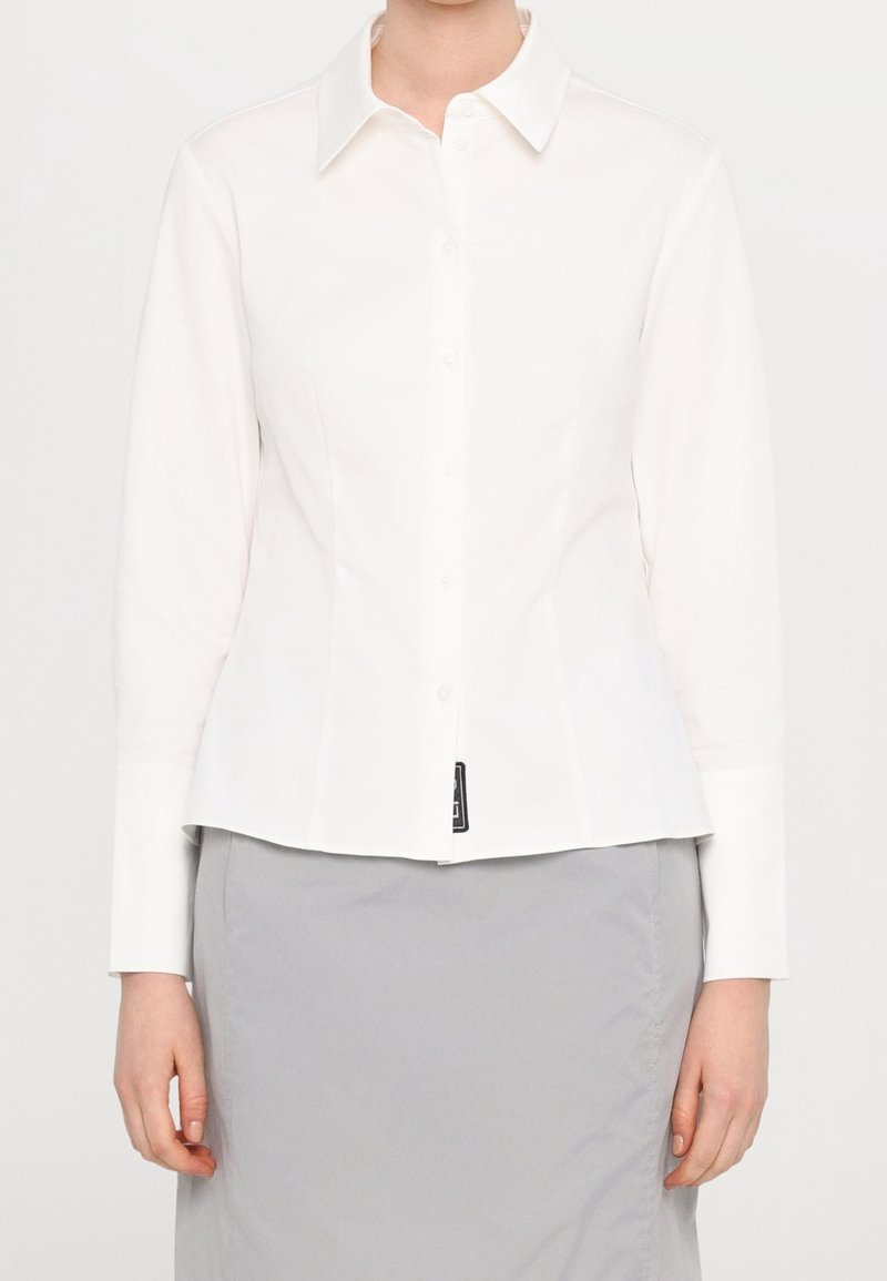Chemise blanche à manches longues avec boutons et col, portée avec une jupe gris clair, montrée du cou aux hanches.