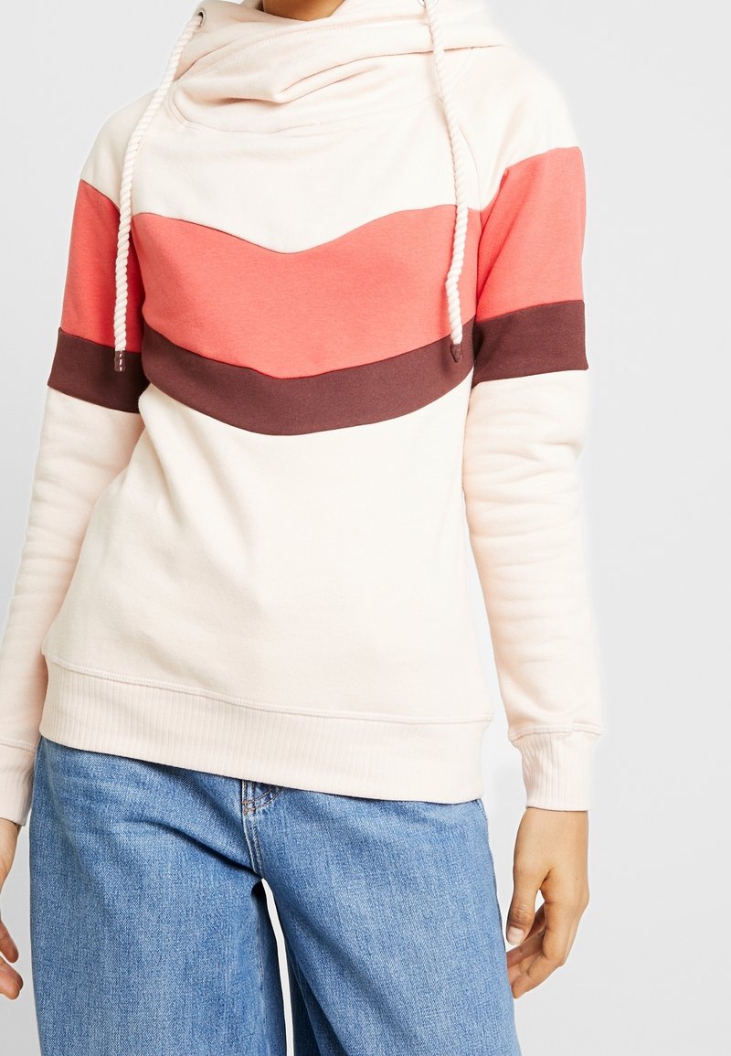 Personne portant un sweat à capuche crème avec un design en blocs de couleur rose et marron et des cordons blancs, associé à un jean bleu clair.