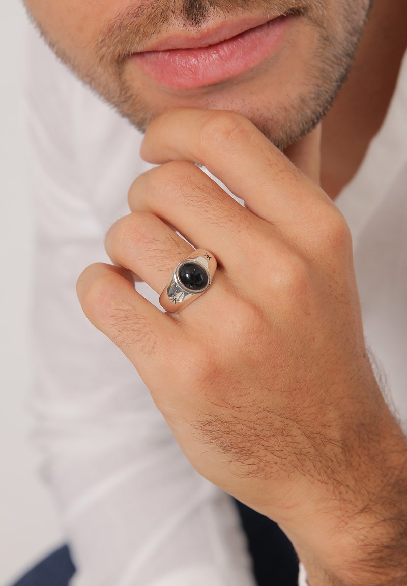 Silverring med en stor svart sten, med släta kanter och en polerad yta. Handen vilar mot ansiktet och visar detaljer.