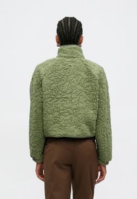 Zelená fleecová bunda s texturou a vysokým límcem, zdobená vzorem listů, s krátkým střihem. Na zadní straně je hladmý lem.