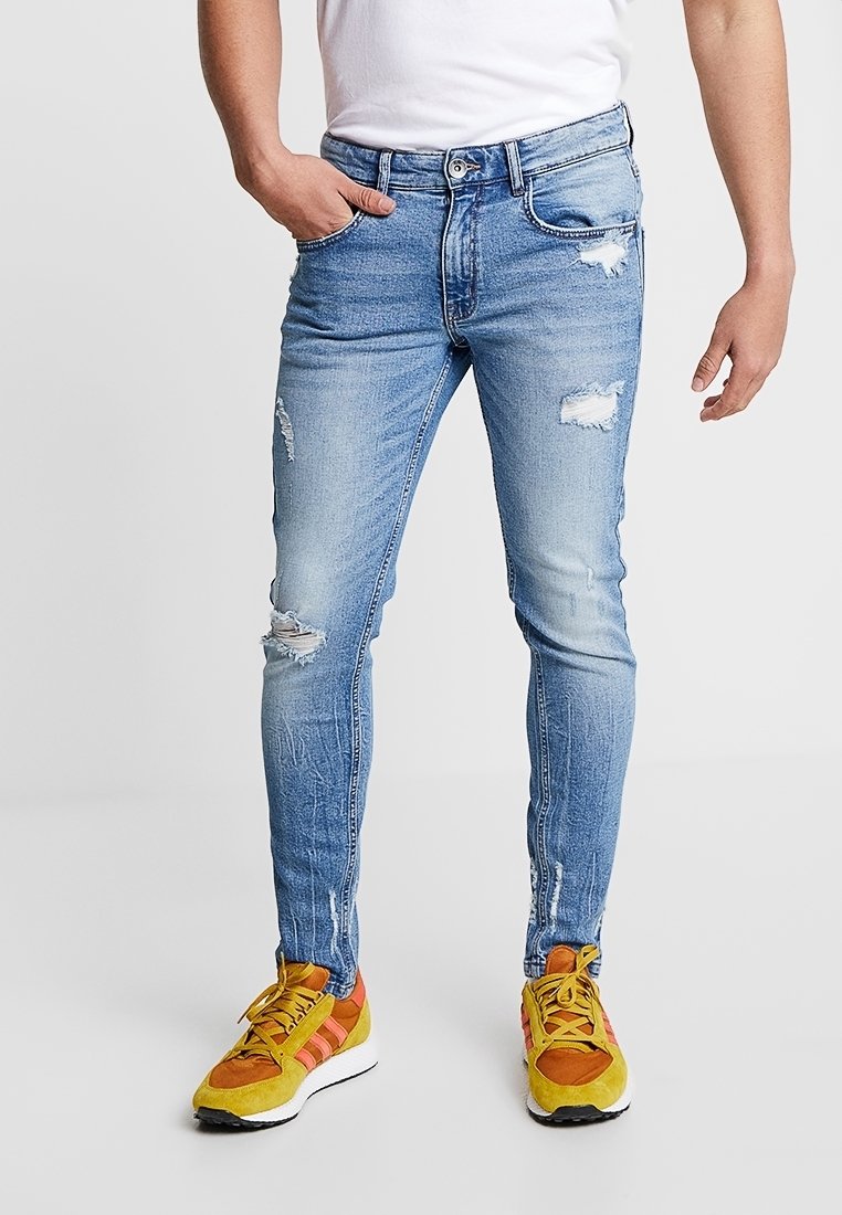 Homme portant un jean skinny déchiré bleu clair et des baskets jaunes et orange, debout avec une main dans la poche sur un fond uni.