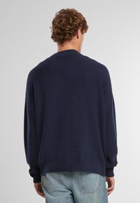 Maglione a maglia blu navy con finitura testurizzata, vestibilità rilassata, spalle scese e polsini a costine. Vista posteriore, mostrato con jeans chiari.