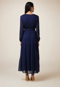Robe maxi bleu marine à manches longues, avec un design à volants, taille cintrée et tissu fluide. Associée à des chaussures à talons jaunes.