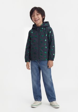 Smiling child wearing navy zip-up jacket with green animal prints, blue jeans, and white sneakers standing against plain background.