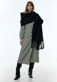 Schwarzer Wollschal, der über ein gemustertes creme- und schwarzfarbenes Kleid drapiert ist. Das Kleid hat einen geometrischen Druck; das Model trägt kniehohe schwarze Stiefel.