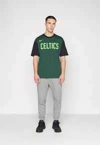 Camiseta bicolor verde y negra con "CELTICS" impreso en letras grandes de color verde lima, combinada con pantalones joggers grises y zapatillas negras.