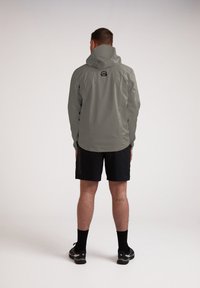Giacca leggera grigia con cappuccio, con un logo visibile. Indossata con pantaloni corti neri e calzini neri, su uno sfondo chiaro e semplice.