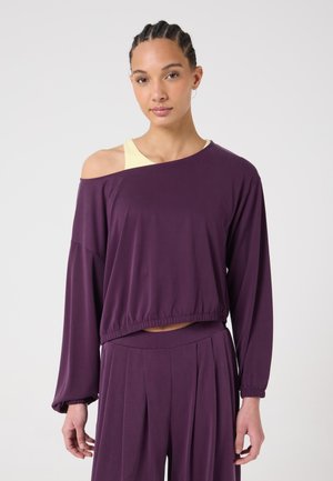 Frau mit geflochtenem Haar trägt ein schulterfreies lila Langarmtop über einem gelben Tanktop und passenden lila Hosen mit weitem Bein.