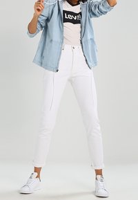 Light blue denim jacket with a hood, black and white graphic Levi's t-shirt, white tapered jeans, and white sneakers. Clean design, casual fit.