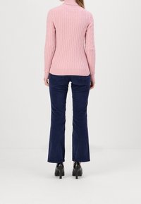 Col roulé en tricot rose clair avec motif torsadé, associé à un pantalon évasé bleu foncé. Des chaussures à talons noires complètent la tenue.