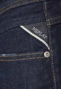 Jeans de mezclilla azul oscuro con una etiqueta de tela blanca y gris que lleva el logo "REPLAY", un botón de latón y detalles de costura en dorado contrastante.
