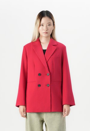 Kvinne med rett, skulderlangt hår iført en oversized rød dobbeltkneppet blazer og lyse bukser foran en enhetlig bakgrunn.