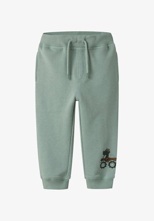 Svijetlo zelene pamučne trenirke s elastikom u struku i vezicom. Sadrži bočne džepove i grafiku traktora na lijevoj nozi.