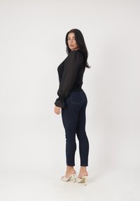 Mujer con cabello largo y oscuro que lleva una blusa negra transparente de manga larga, vaqueros ajustados oscuros y sandalias beige de tacón alto, de pie de lado.
