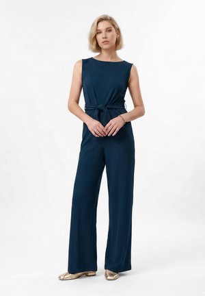 Frau mit kurzen blonden Haaren, die einen ärmellosen dunkelblauen Overall trägt, der in der Taille gebunden ist, und goldene flache Schuhe. Sie steht mit verschränkten Händen.