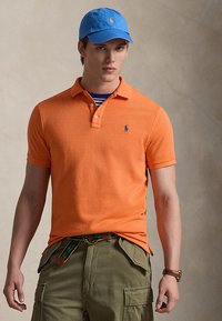 Noor mees, kes kannab sinist mütsi, apelsinivärvilist polo-särki väikese logoga, triibulist alussärki, oliivrohelisi cargo-pükse ja pruuni kella, seisab neutraalse tausta ees.