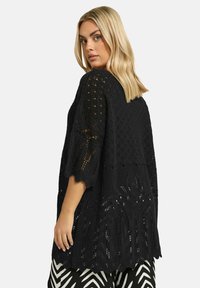 Mustat virkattuja neuletakit, joissa on leveät hihat, tekstuurinen kuvio ja aaltoileva helma, yhdistettynä mustavalkoisiin kuviollisiin housuihin.