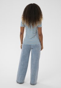 Gestreiftes, tailliertes Oberteil in Blau und Weiß mit wellenförmigen Kanten, kombiniert mit hellblauen Jeans mit weitem Bein. Haare in lockeren, voluminösen Locken gestylt.