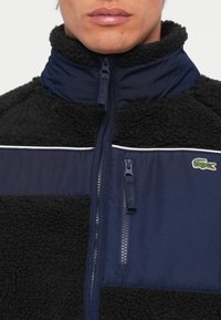 Schwarze Fleecejacke mit navyblauen Nylonakzenten, hohem Kragen, einer Reißverschlusstasche und einem kleinen grünen Krokodil-Logo auf der Brust.
