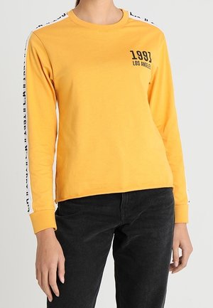 Personne portant une chemise à manches longues jaune moutarde avec le texte "1993 Los Angeles" et une bande blanche avec des lettres noires sur les manches, associée à un pantalon noir.