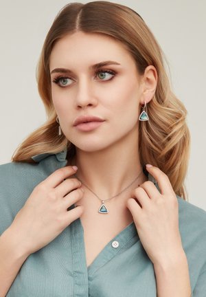 Femme aux cheveux châtain clair portant un collier et des boucles d'oreilles assortis en forme de pierre précieuse triangulaire bleue, vêtue d'une chemise boutonnée bleu sarcelle, regardant sur le côté.