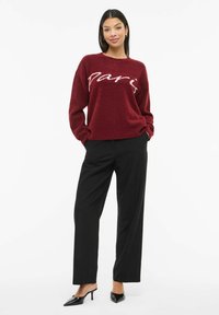 Bordeauxrode gebreide trui met witte "Paris" tekst, losse pasvorm, ronde hals, gecombineerd met zwarte wijde pijpen broek en puntige zwarte hakken.