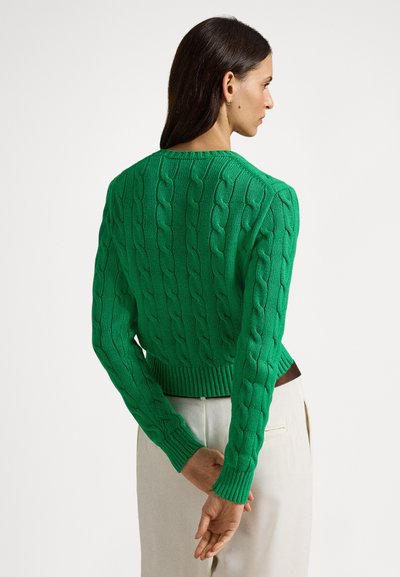 Camisola verde tricotada com padrões de trança, decote redondo e punhos e barra canelados. Modelo de costas para a câmara, vestindo calças creme.