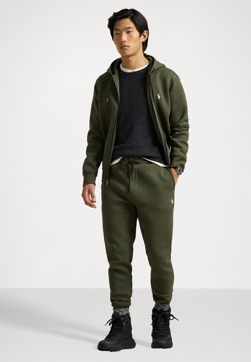 Sudadera verde con cremallera, suéter gris debajo, pantalones de chándal verdes a juego y botas negras. Las características notables incluyen un logo en el pecho.