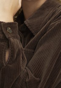 Bruine corduroy shirt met een klassieke kraag, twee knopen op de manchetten en een gemêleerd ribbelpatroon. Close-up focus op stof en ontwerp.