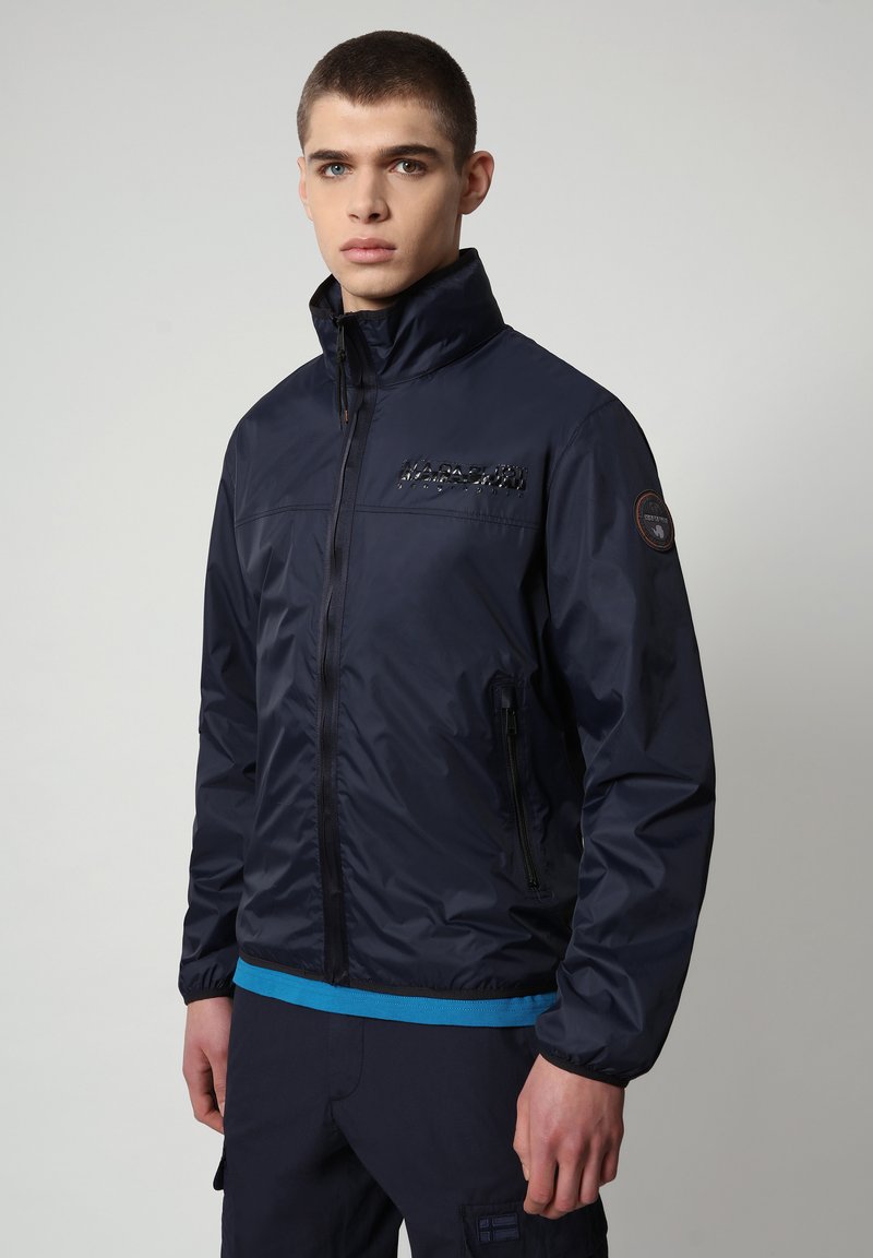 Giacca impermeabile blu navy con colletto alto, chiusura frontale con zip e logo nero sul petto; presenta rifiniture blu a contrasto e tasche con zip.