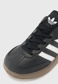 Musta urheilukenkä, jossa on teksturoitu nahkainen päällinen, mustat nauhat ja valkoiset raidat. Kenkässä on ruskea kumipohja ja musta logo kielen päällä.