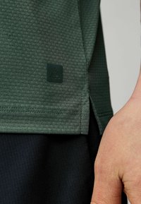 Grøn tekstureret atletisk skjorte med en lille sort logo detalje på kanten, featuring et sidesplit og tætsiddende design.