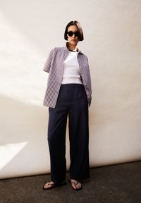 Kortärmad rutiga skjorta i lila och vitt, lager över en vit topp, kombinerat med vida marinblå byxor och svarta flip-flops.