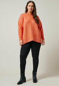 Oranges strukturierter Pullover mit hohem Kragen, diagonalem Rippmuster und abgerundetem Saum, kombiniert mit engen schwarzen Hosen und Stiefeln mit Absatz.