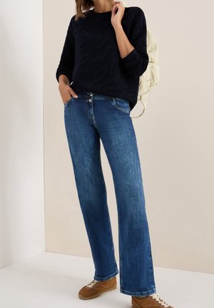 Femme portant un haut à manches longues bleu foncé, un jean droit bleu, des baskets marron, et un sac à dos matelassé beige clair sur une épaule.